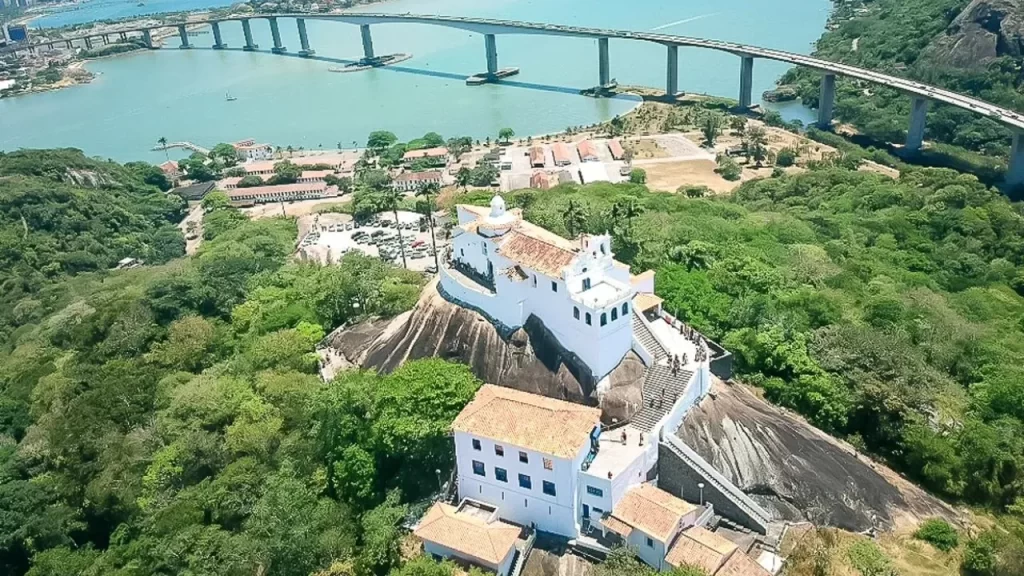 Explore o Espírito Santo: Os 10 Destinos Mais Procurados que Você Precisa Conhecer 1 Vista aérea panorâmica do Convento da Penha, um edifício branco histórico no topo de um morro verde, com a imponente Terceira Ponte atravessando a baía azul clara. Ao fundo, a orla da cidade de Vila Velha e parte de Vitória, no Espírito Santo, sob um céu ensolarado.
