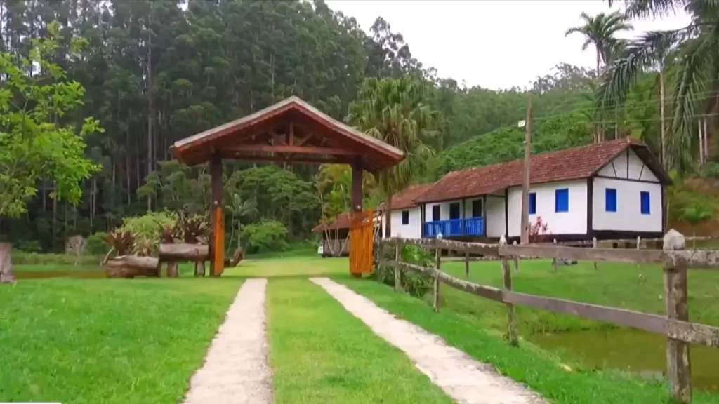 Rotas Turísticas no Espírito Santo: Os Melhores Destinos para Explorar o Estado 2 Rota dos Vinhos: Caminho de grama verde levando a um portão de madeira rústico coberto, marcando a entrada de uma propriedade rural em Guarapari. Uma casa branca com detalhes azuis e telhado de barro é visível ao fundo, cercada por vegetação densa, incluindo árvores altas e palmeiras. Uma cerca de madeira acompanha o caminho.