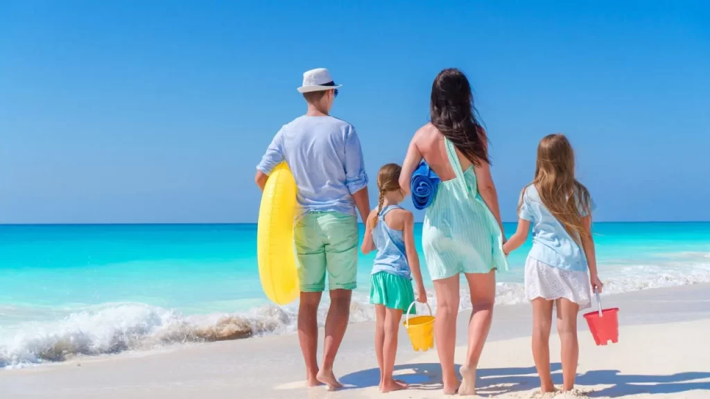 Descubra os Destinos Mais Procurados para Viajar no Verão 2025! 1 os Destinos Mais Procurados para Viajar: Uma família de quatro pessoas, vista de costas, caminhando juntas na areia clara de uma praia ensolarada em Guarapari, com o mar azul turquesa com pequenas ondas ao fundo. O pai segura uma boia amarela e a mãe uma toalha azul enrolada. As duas filhas, de cabelos longos, andam de mãos dadas com os pais. Uma menina carrega um balde amarelo e a outra um balde vermelho. O céu está claro e azul.