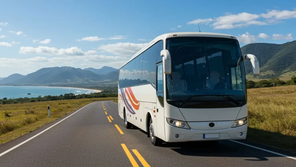 Reivax Viagens: Descubra o Brasil com Conforto, Segurança e Experiência 3 Ônibus branco moderno viajando por uma estrada asfaltada com duas faixas amarelas, margeada por vegetação rasteira e com o oceano, montanhas e céu azul com nuvens ao fundo.