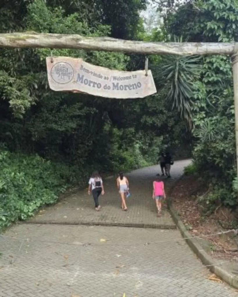 Um arco rústico de madeira com a placa "Bem-vindo ao Morro do Moreno" sobre um caminho pavimentado que leva a uma área verde, com pessoas caminhando.