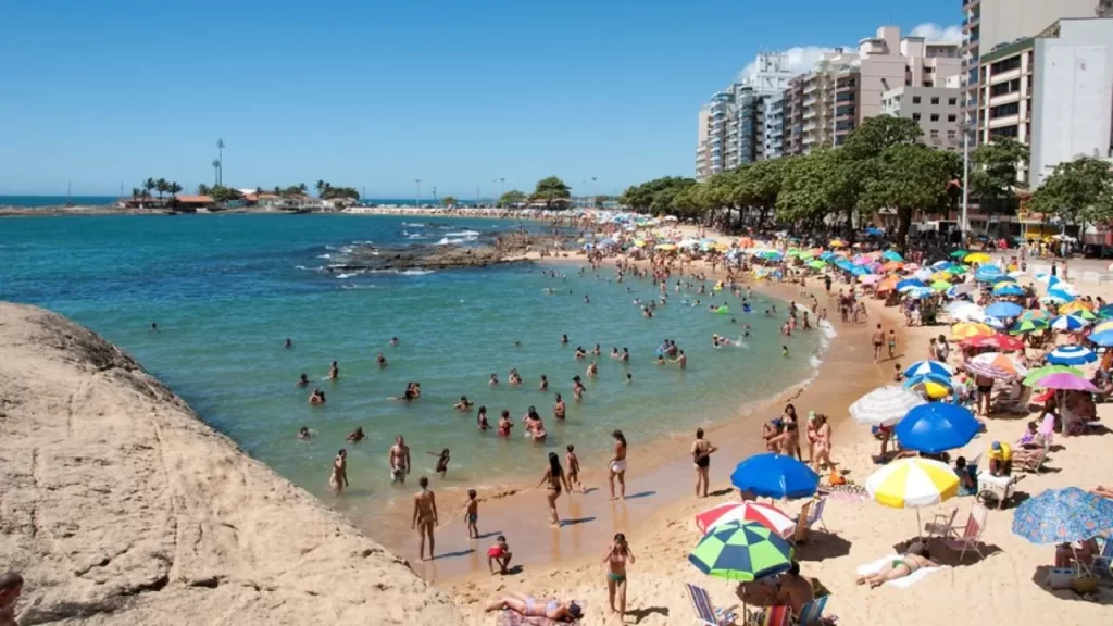 O Litoral Capixaba em Alta: Guarapari Lidera a Busca por Viagens de Bem-Estar e Família 3 Vista da Praia da Areia Preta em Guarapari, mostrando uma grande quantidade de pessoas aproveitando o dia ensolarado. Há muitos guarda-sóis coloridos na areia e diversas pessoas nadando e se divertindo na água. Edifícios altos margeiam a praia ao fundo.