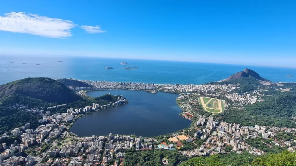 Descubra os Destinos Mais Procurados para Viajar no Verão 2025! 2 Destinos Mais Procurados para Viajar: Uma vista aérea ampla de Guarapari, mostrando uma grande lagoa de água escura cercada por uma densa área urbana de edifícios baixos e vegetação. Montanhas verdejantes delimitam a cidade, e ao fundo, o oceano azul encontra a costa com praias de areia clara. O céu está claro e azul com algumas nuvens brancas. Uma pista de corrida oval pode ser vista perto da lagoa.