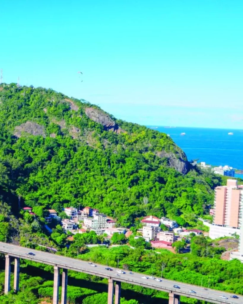 Uma imagem de ângulo alto mostrando o verde Morro do Moreno, um parapente no céu, a cidade abaixo com vários edifícios e a Segunda Ponte se estendendo sobre a água à distância.