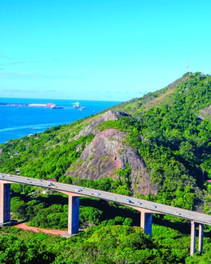 Uma vista panorâmica das encostas verdes e rochosas do Morro do Moreno à direita, de frente para o oceano azul com navios e a Segunda Ponte elevada com carros em movimento em primeiro plano.