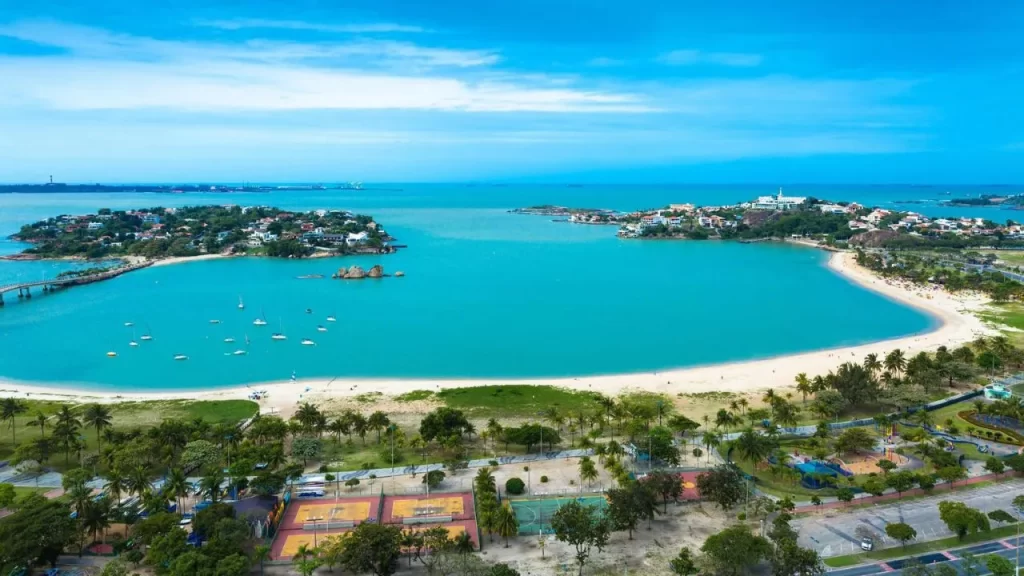 O Litoral Capixaba em Alta: Guarapari Lidera a Busca por Viagens de Bem-Estar e Família 1 Litoral Capixaba em Alta: Ampla vista aérea da Baía do Mucugê em Guarapari, com águas azul-turquesa, praias de areia clara, vegetação costeira exuberante e barcos navegando na baía. Ao fundo, vislumbra-se o mar aberto sob um céu azul com poucas nuvens.