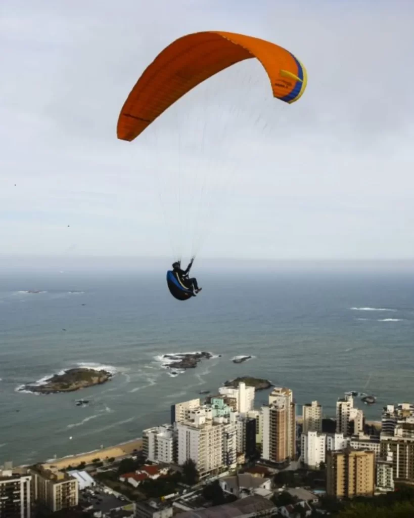 Um parapente com uma vela laranja e azul sobrevoa a cidade de Vila Velha, com o oceano e ilhas pequenas abaixo e o Morro do Moreno visível no horizonte à direita