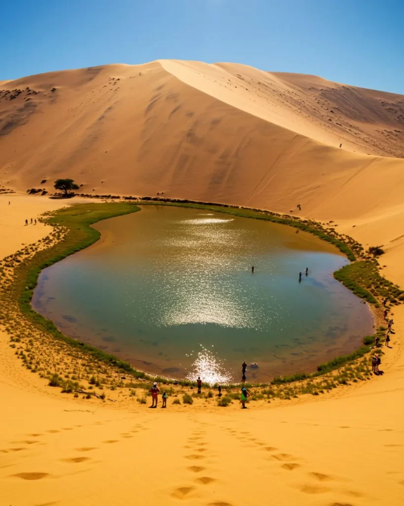 Vista de cima de um oásis de água esverdeada cercado por vegetação rasteira no meio de vastas dunas de areia laranja sob um céu azul claro, com pequenos grupos de pessoas caminhando e se refrescando na água. WayVix Viagens.
