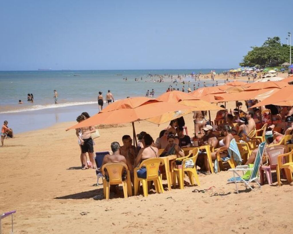 Balneabilidade nas Praias da Serra: Guia Atualizado para Banho Seguro 1 Balneabilidade nas Praias da Serra: Pessoas relaxando sob grandes guarda-sóis cor de laranja em uma praia ensolarada. A areia está cheia de cadeiras e mesas de plástico, com grupos de amigos e famílias aproveitando a tarde. No fundo, o mar está calmo e há mais pessoas nadando e caminhando pela orla.