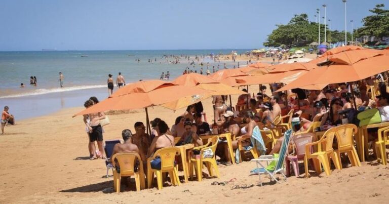 Pessoas relaxando sob grandes guarda-sóis cor de laranja em uma praia ensolarada. A areia está cheia de cadeiras e mesas de plástico, com grupos de amigos e famílias aproveitando a tarde. No fundo, o mar está calmo e há mais pessoas nadando e caminhando pela orla.