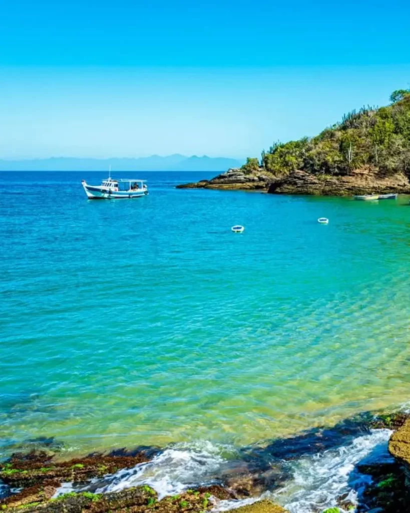 Búzios, RJ: o guia completo para encontrar o refúgio ideal com vista para o mar 3 Uma vista da praia de Búzios, RJ, com água azul-turquesa, algumas embarcações flutuando e uma costa rochosa.