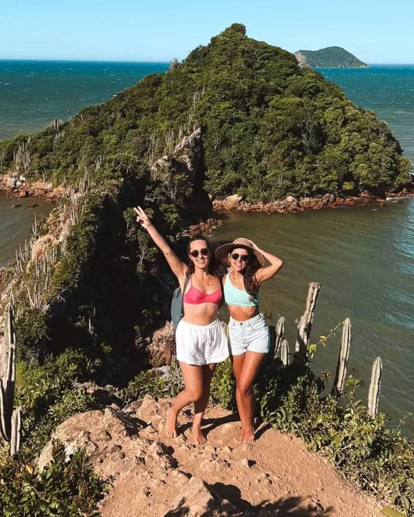 Búzios, RJ: o guia completo para encontrar o refúgio ideal com vista para o mar 2 Duas mulheres em um penhasco rochoso, com o mar e uma ilha coberta de vegetação ao fundo, em Búzios, RJ
