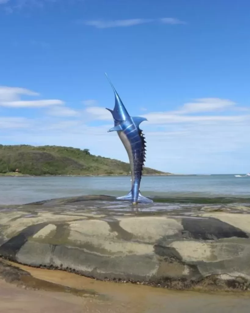 Guarapari em 3 Dias: Estátua do peixe-agulha, um dos símbolos de Guarapari, com o mar e o morro ao fundo. Um ponto de referência para quem visita a cidade em um roteiro completo.