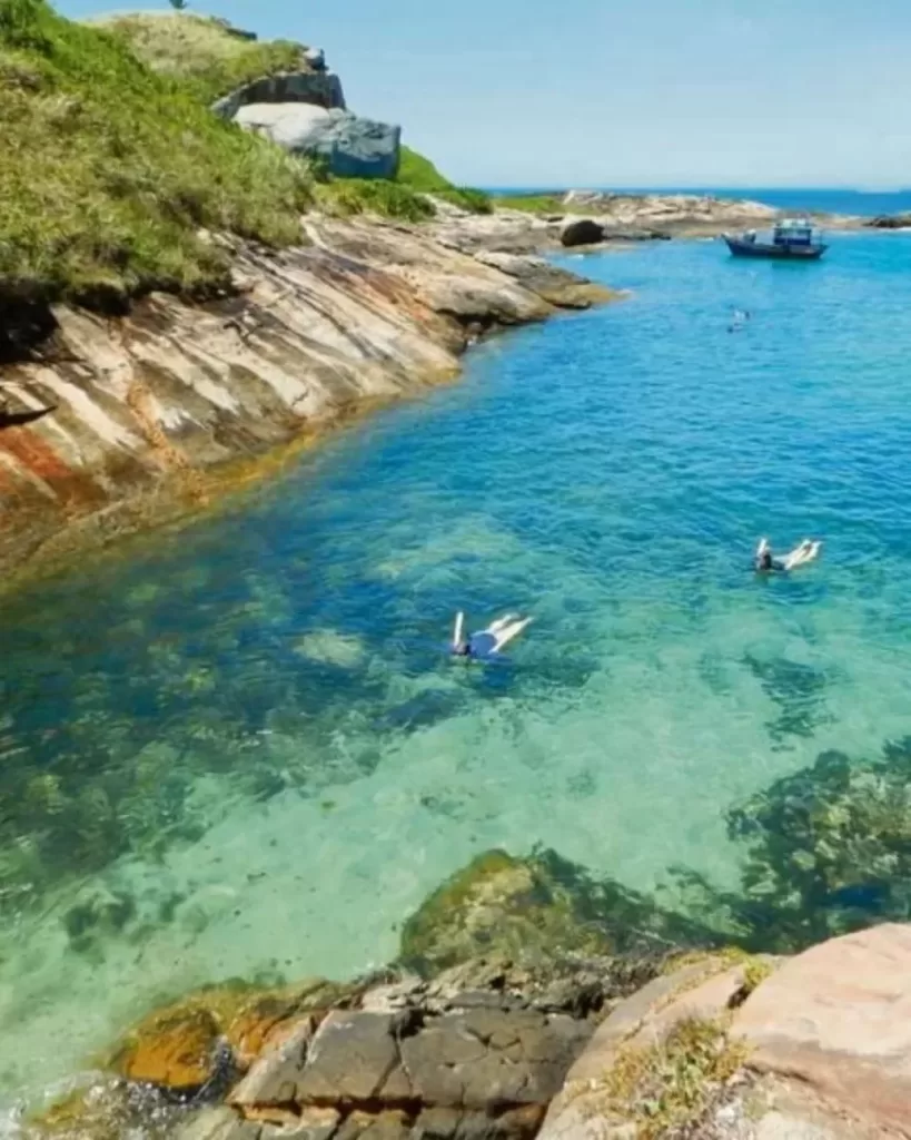 Guarapari em 3 Dias: Pessoas mergulhando em águas claras e azuis de uma enseada em Guarapari, cercada por rochas e vegetação. Uma atividade imperdível no roteiro de 3 dias para explorar as belezas naturais da cidade.