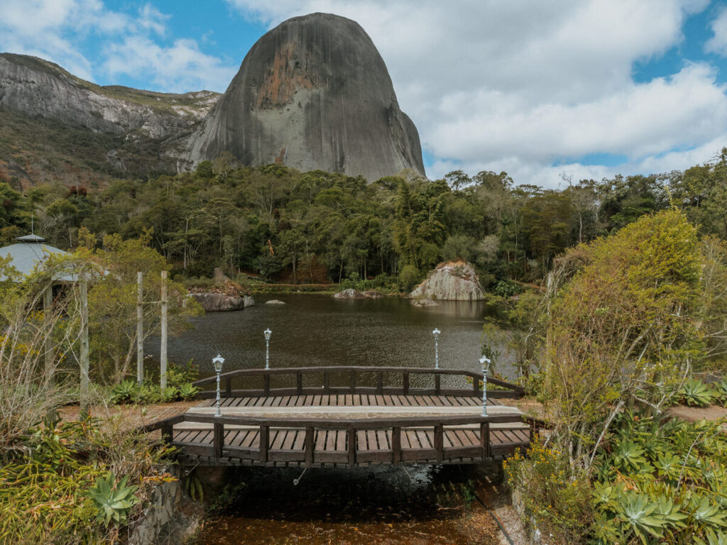 Pedra Azul: Guia completo de trilhas e piscinas naturais 3 O que fazer na Pedra Azul: A imponente Pedra Azul em destaque, vista à distância de um lago calmo. Em primeiro plano, uma pequena ponte de madeira com grades e postes de luz. A paisagem é cercada por vegetação exuberante e árvores.