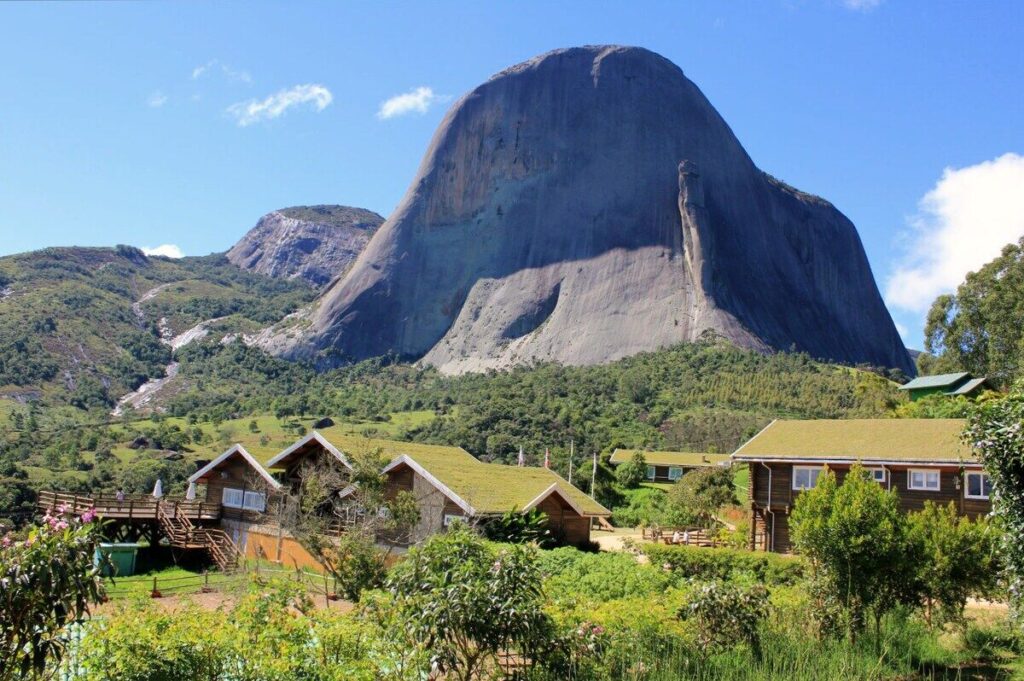 Pedra Azul: Guia completo de trilhas e piscinas naturais 2 Chalés com telhados verdes e arquitetura rústica, cercados por vegetação, com a imponente Pedra Azul e as montanhas ao fundo. O céu está claro e parcialmente nublado.