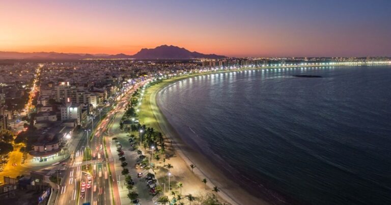 Vista da Praia da Costa ao entardecer, com o sol se pondo e cores quentes no céu, pessoas na areia e no mar, e prédios ao fundo.