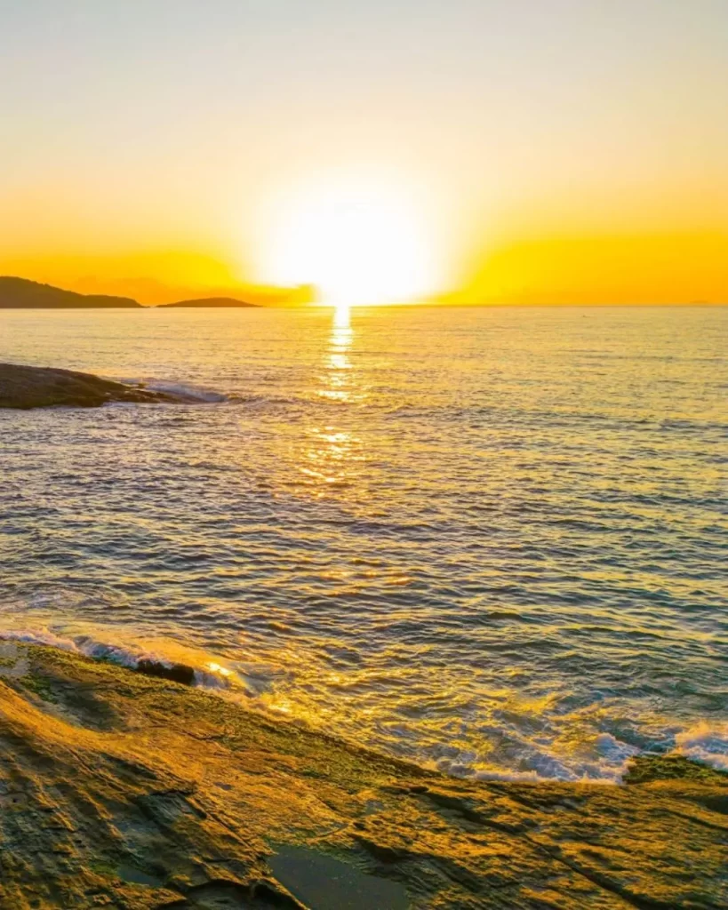 Melhores praias do Espírito Santo: guia completo para quem quer explorar o litoral capixaba em 2025 3 Um belo pôr do sol sobre o mar, com o sol em um tom dourado e brilhante refletindo na água e nas rochas da costa. Esta cena de fim de tarde destaca a tranquilidade e a beleza natural que fazem de Guarapari uma das 10 melhores praias do Espírito Santo para se visitar.