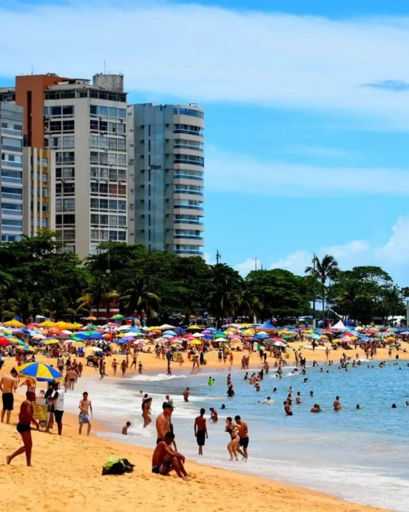 Melhores praias do Espírito Santo: guia completo para quem quer explorar o litoral capixaba em 2025 1 Uma vista panorâmica da Praia da Costa em Vila Velha, com prédios altos ao fundo, coqueiros, e a areia cheia de guarda-sóis coloridos e pessoas aproveitando o sol. A água do mar é calma e muitas pessoas estão nadando, mostrando por que é considerada uma das 10 melhores praias do Espírito Santo.