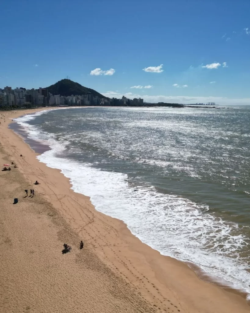 Melhores praias do Espírito Santo: guia completo para quem quer explorar o litoral capixaba em 2025 2 Uma vista aérea da Praia de Camburi em Vitória, com a orla e o calçadão, onde a areia dourada se encontra com as ondas do mar. Ao fundo, pode-se ver o Morro do Moreno e a cidade, destacando a beleza que a coloca na lista das 10 melhores praias do Espírito Santo.