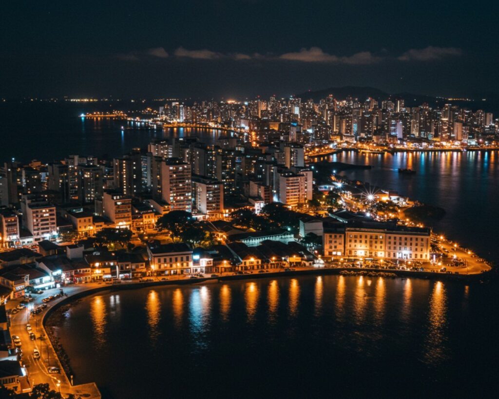 Vida Noturna em Vitória: Um Guia Completo para Bares e Restaurantes 2 vida noturna em Vitória: Uma vista aérea e panorâmica da cidade de Vitória, Espírito Santo, à noite. A foto mostra a baía de águas escuras refletindo as luzes douradas e brilhantes dos edifícios. As construções do centro histórico se destacam em primeiro plano, enquanto a silhueta da cidade moderna e das colinas brilha no fundo.