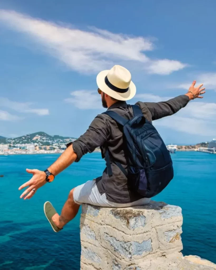 Como funciona o RCI viagens? Guia completo para transformar sua propriedade de férias em experiências incríveis 4 Um viajante de chapéu e mochila sentado em um muro de pedra, com os braços abertos, admirando a vista do mar e de uma cidade costeira. Entenda como funciona o RCI viagens para explorar novos horizontes.