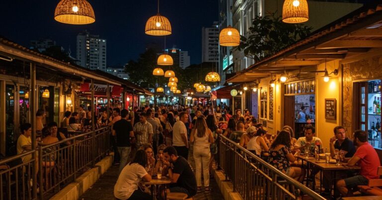 Foto de uma rua movimentada à noite, repleta de pessoas em mesas de bares e restaurantes. O ambiente é iluminado por lâmpadas penduradas e as calçadas estão cheias de pedestres, capturando a energia da vida noturna de Vitória, Espírito Santo.